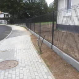 Nowoczesne ogrodzenie panelowe w kolorze antracytowym z podmurówką betonową, widoczne z perspektywy chodnika z kostki brukowej w otoczeniu zieleni i zabudowań.