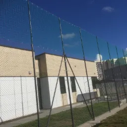 Ochronna siatka ogrodzeniowa z drobnymi oczkami na metalowych słupkach, zabezpieczająca teren boiska sportowego przy budynku z cegły, widok w słoneczny dzień.