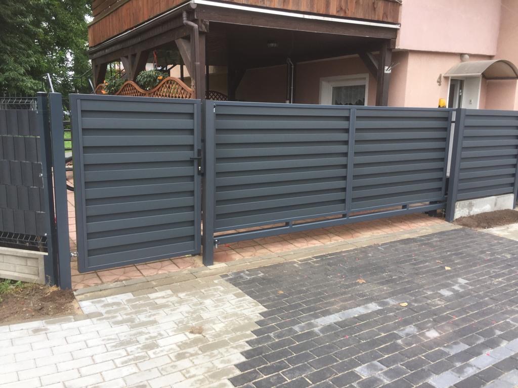 Nowoczesna, szara brama wjazdowa z poziomymi panelami, częściowo otwarta, z widocznym fragmentem ogrodzenia panelowego i kostką brukową przed domem.