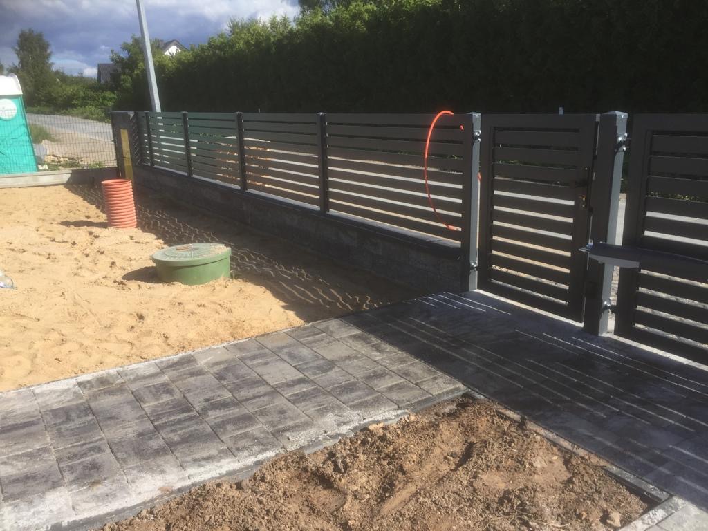 Nowoczesne, poziome panele ogrodzeniowe w kolorze grafitowym z bramą wjazdową, widoczne elementy montażowe, świeżo ułożona kostka brukowa, teren w trakcie zagospodarowywania.