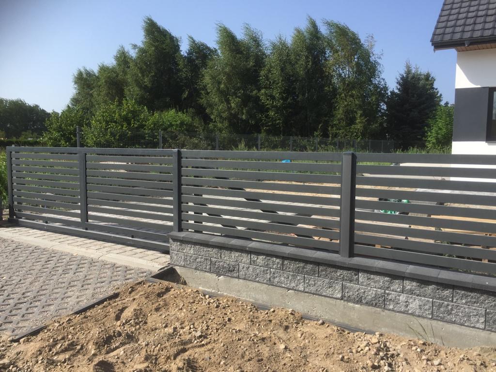 Nowoczesna, pozioma brama przesuwna z metalowymi lamelami w kolorze grafitowym, zamontowana na murku z bloczków betonowych, z widocznym podjazdem z kostki brukowej i nasypanym piaskiem.