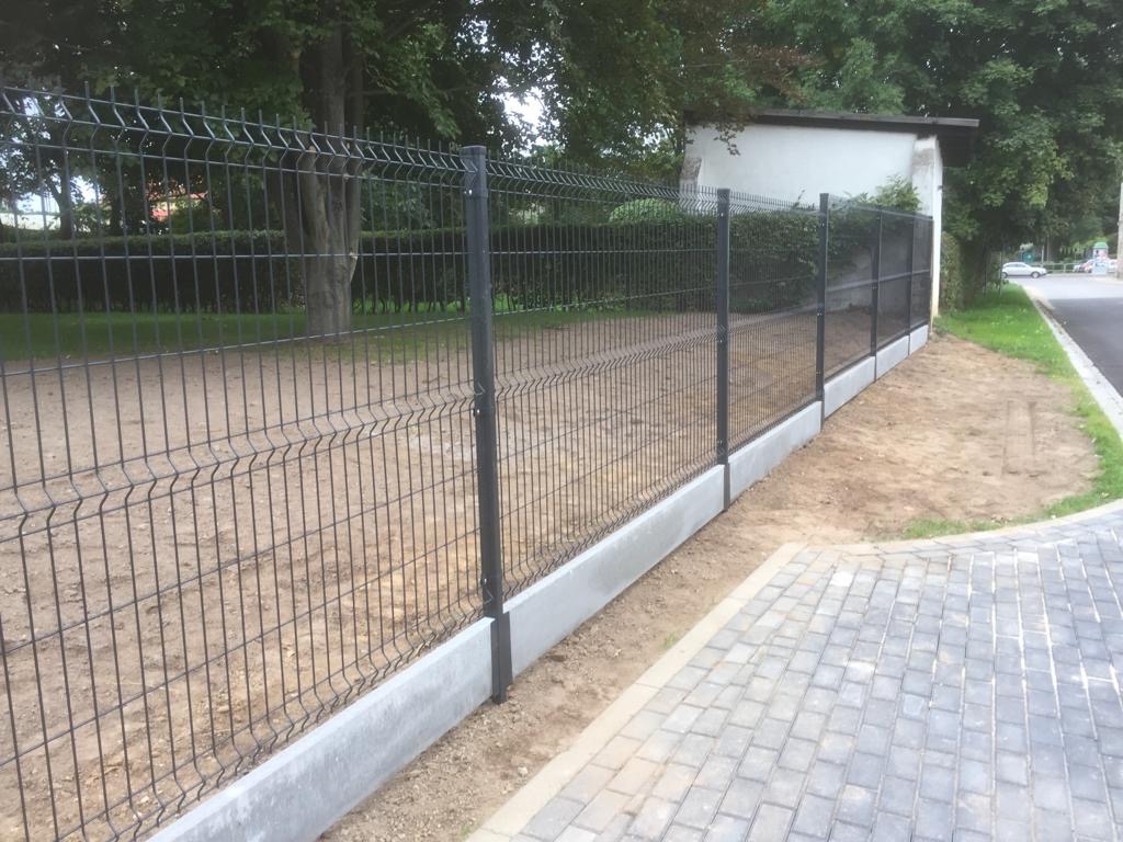 Ciemnoszare ogrodzenie panelowe z podmurówką betonową, widok z boku, kostka brukowa na pierwszym planie, trawa i drzewa w tle.