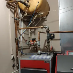 Używany, lekko zakurzony piec na paliwo stałe z instalacją hydrauliczną i zbiornikiem wyrównawczym w kotłowni.