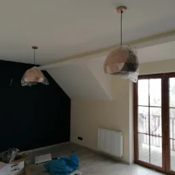 Wnętrze poddasza po malowaniu ścian na czarno i beżowo, z zamontowanymi, ale jeszcze ofoliowanymi lampami w geometryczne wzory, widoczny grzejnik i okno balkonowe.