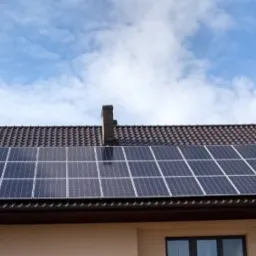 Panele słoneczne zamontowane na brązowym dachu domu jednorodzinnego z widocznym kominem i błękitnym niebem w tle.