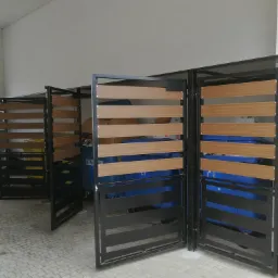 Zadaszone, modułowe osłony śmietnikowe z poziomych, drewnopodobnych paneli i czarnej ramy, chroniące przed wiatrem i wzrokiem.