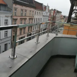 Nierdzewna balustrada balkonowa z widokiem na starą zabudowę miejską, mocowana na metalowych słupkach przykręcanych śrubami.