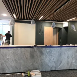 Pracownik maluje ścianę na drabinie w biurze z marmurową ladą, zakrytymi lampami i drewnianym sufitem lamelowym.