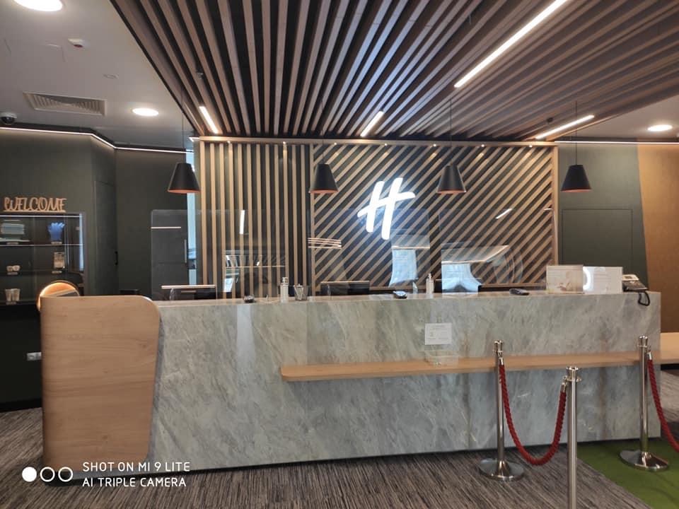 Recepcja w nowoczesnym biurze z marmurowym blatem, drewnianą zabudową, oświetleniem punktowym i dekoracyjnym sufitem z drewnianych listew.