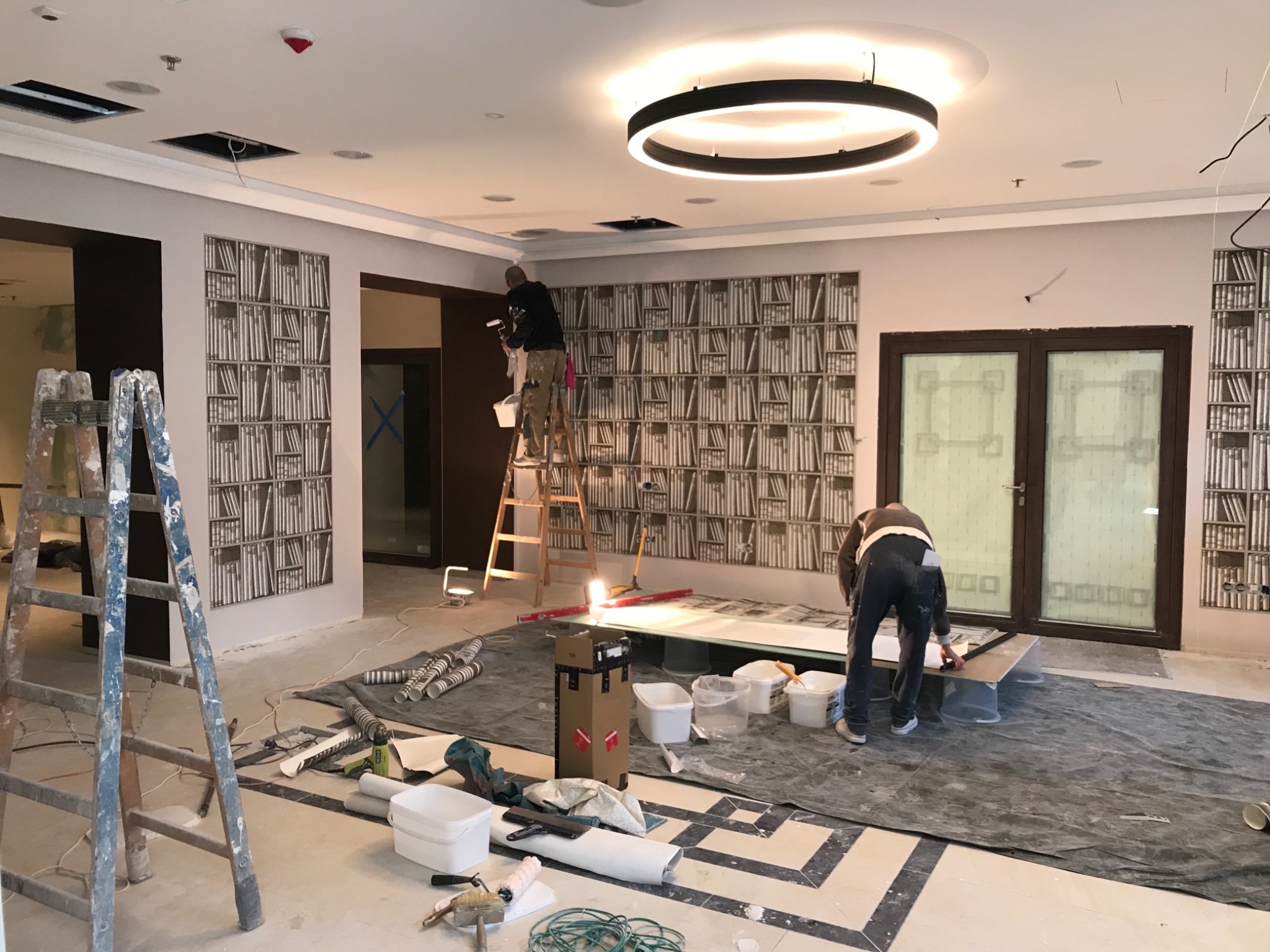 Dwóch pracowników podczas prac wykończeniowych w pomieszczeniu z tapetą imitującą regał z książkami; jeden na drabinie maluje sufit, drugi przygotowuje arkusz tapety na podłodze zabezpieczonej folią.