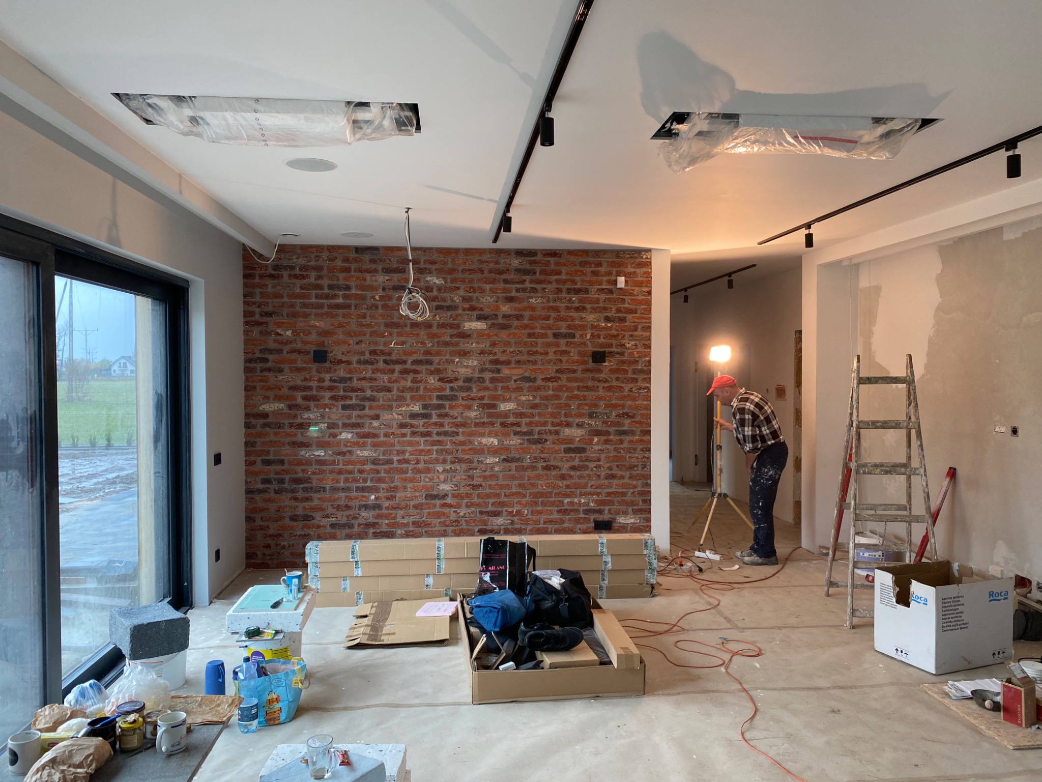 Wnętrze w trakcie prac wykończeniowych: ściana z cegły, surowy beton, oświetlenie szynowe, drabina i pracownik z lampą na statywie. Widoczne materiały budowlane i narzędzia.