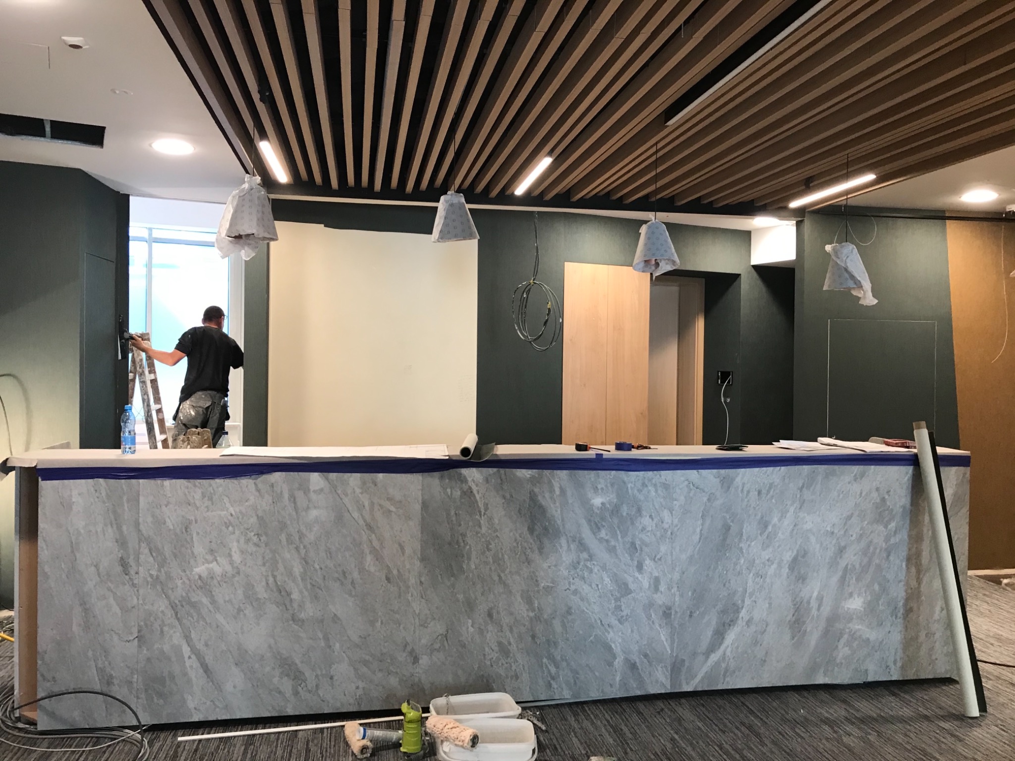 Pracownik maluje ścianę na drabinie w biurze z marmurową ladą, zakrytymi lampami i drewnianym sufitem lamelowym.