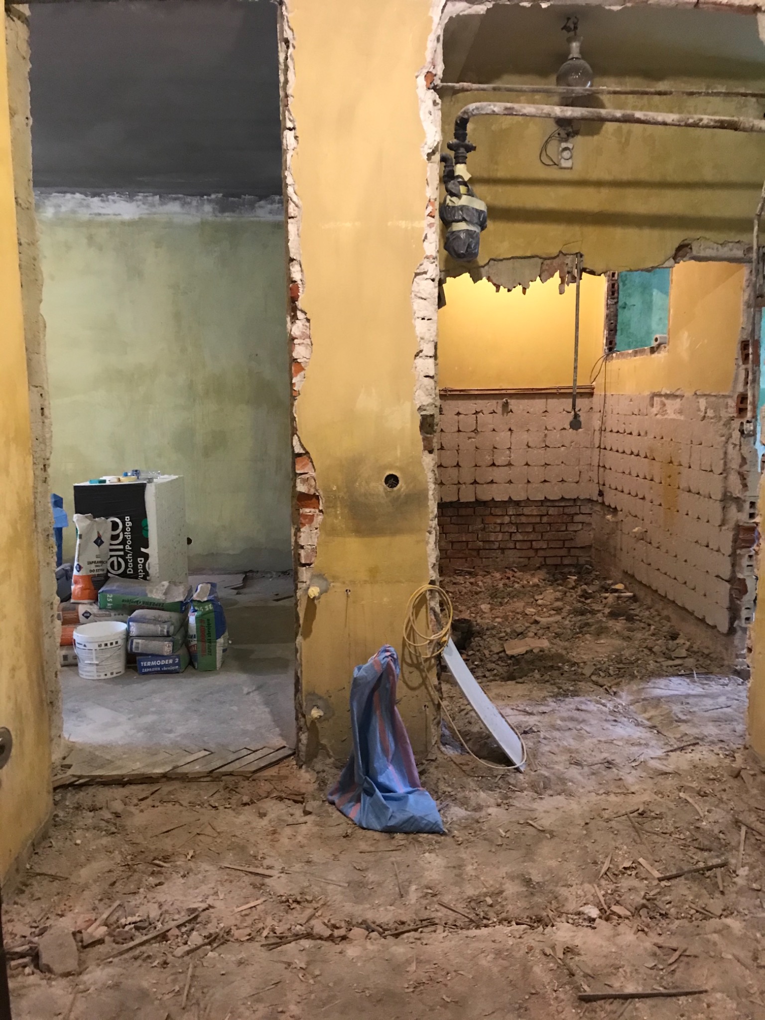 Wnętrze mieszkania w trakcie generalnego remontu, widoczne odsłonięte cegły, gruz na podłodze, worki z materiałami budowlanymi i instalacje elektryczne.