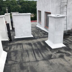 redlabpro specjalistyczne usługi remontowo budowlane - Świeżo pomalowane na biało kominy wentylacyjne na czarnym, płaskim dachu budynku po renowacji, z widocznymi śladami farby i drabinką ewakuacyjną na bocznej ścianie.