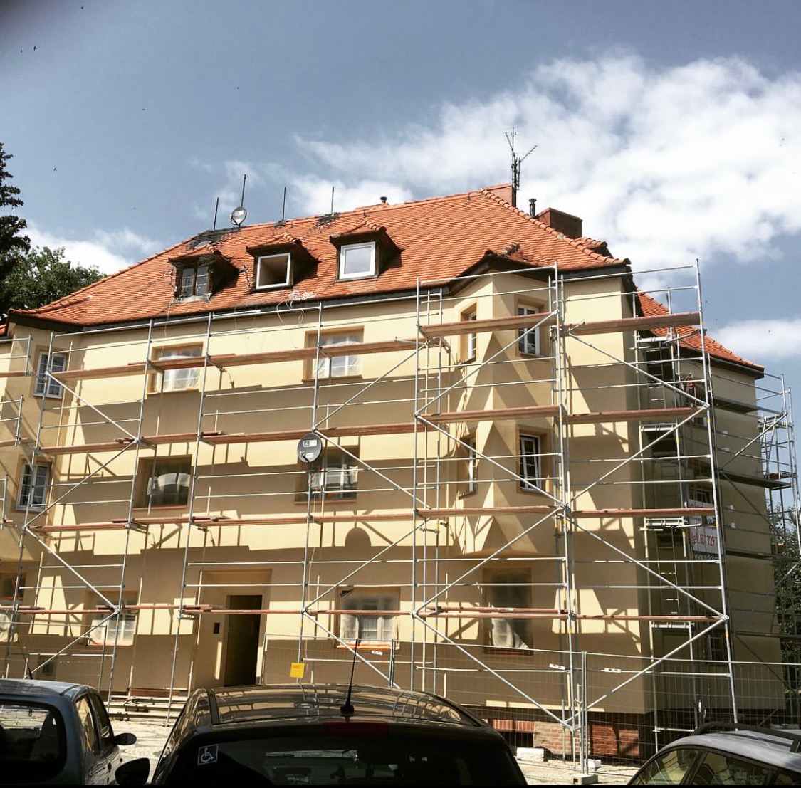 Żółty budynek mieszkalny w trakcie prac elewacyjnych, otoczony rusztowaniem, z widoczną nową czerwoną dachówką i anteną na dachu.
