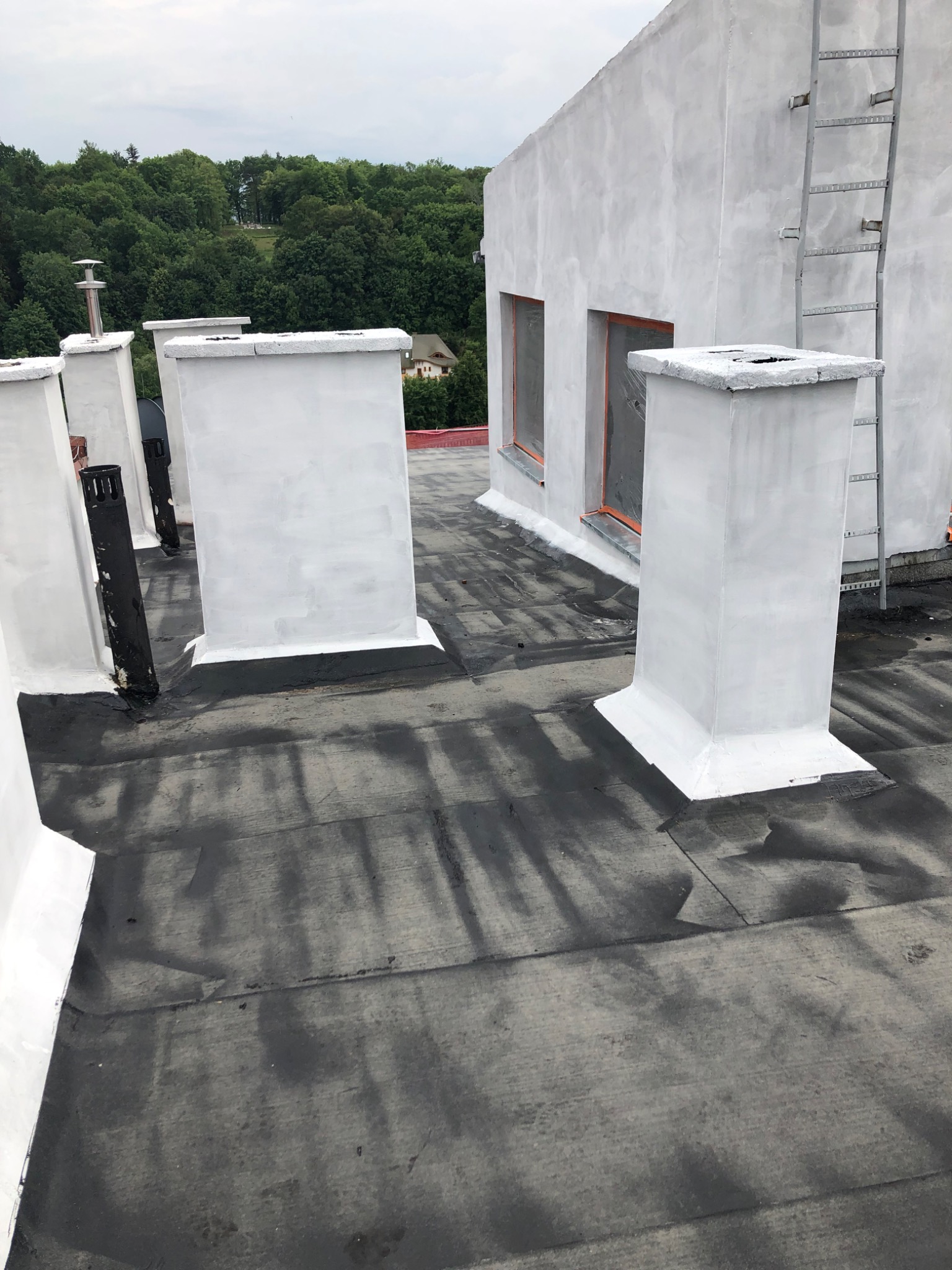 Świeżo pomalowane na biało kominy wentylacyjne na czarnym, płaskim dachu budynku po renowacji, z widocznymi śladami farby i drabinką ewakuacyjną na bocznej ścianie.