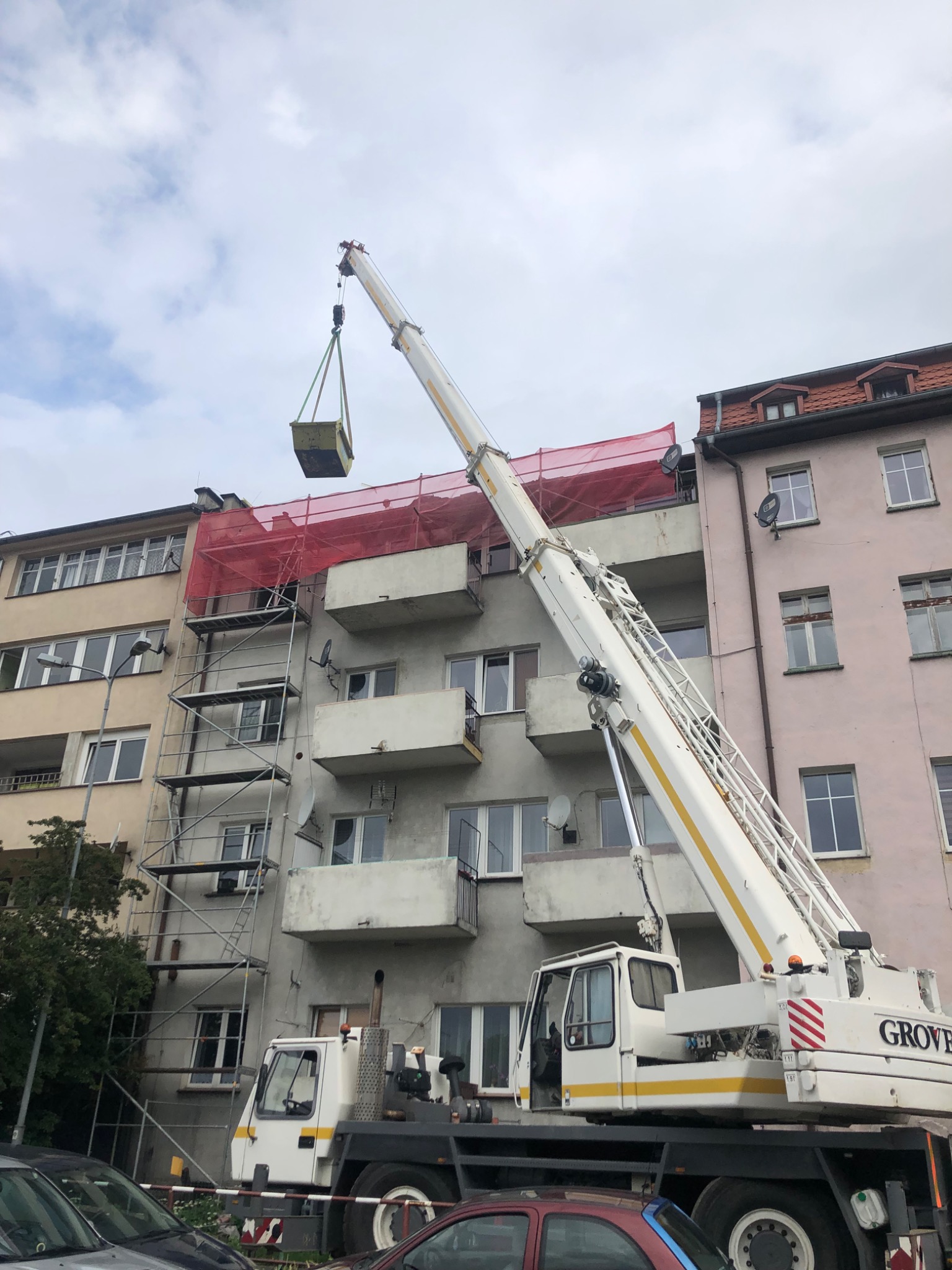 Żuraw podnosi kontener na rusztowanie przed budynkiem mieszkalnym z balkonami, zabezpieczonym czerwoną siatką.