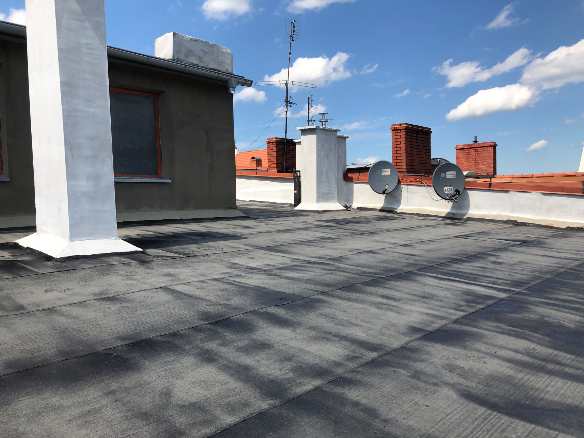 Widok na płaski dach pokryty papą termozgrzewalną z kominami i antenami satelitarnymi w słoneczny dzień.