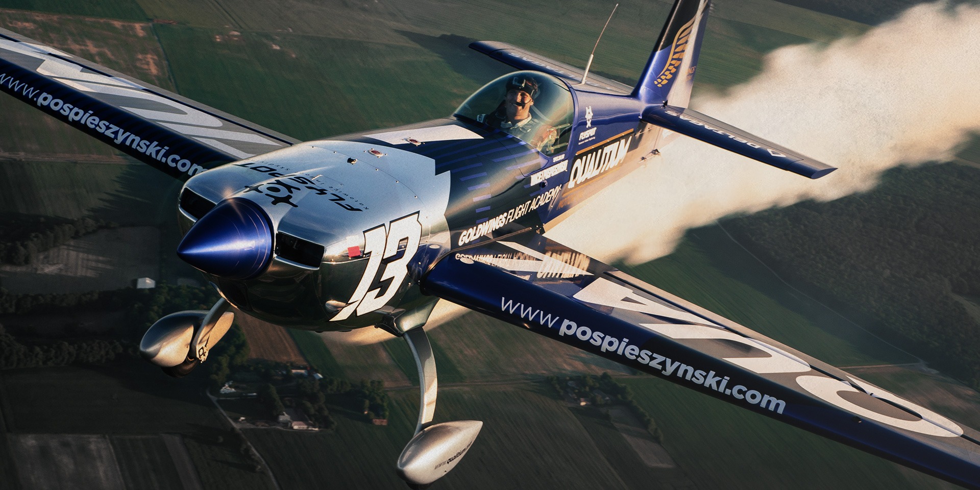 Akrobacyjny samolot w locie, pilot uśmiechnięty w kokpicie, widok z góry na zielone pola, oznaczenia firmowe na skrzydłach, dym za samolotem.