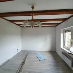 Pusty pok&oacute;j po odświeżeniu z drewnianymi belkami na suficie, nową podłogą i świeżo pomalowanymi ścianami na biało, widoczny grzejnik pod oknem i elementy montażowe na podłodze.