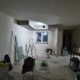 Wnętrze w trakcie remontu: widoczne płyty karton gips, drabina, narzędzia, zakryte folią okno i świeżo wygładzone ściany pod przyszłe malowanie.