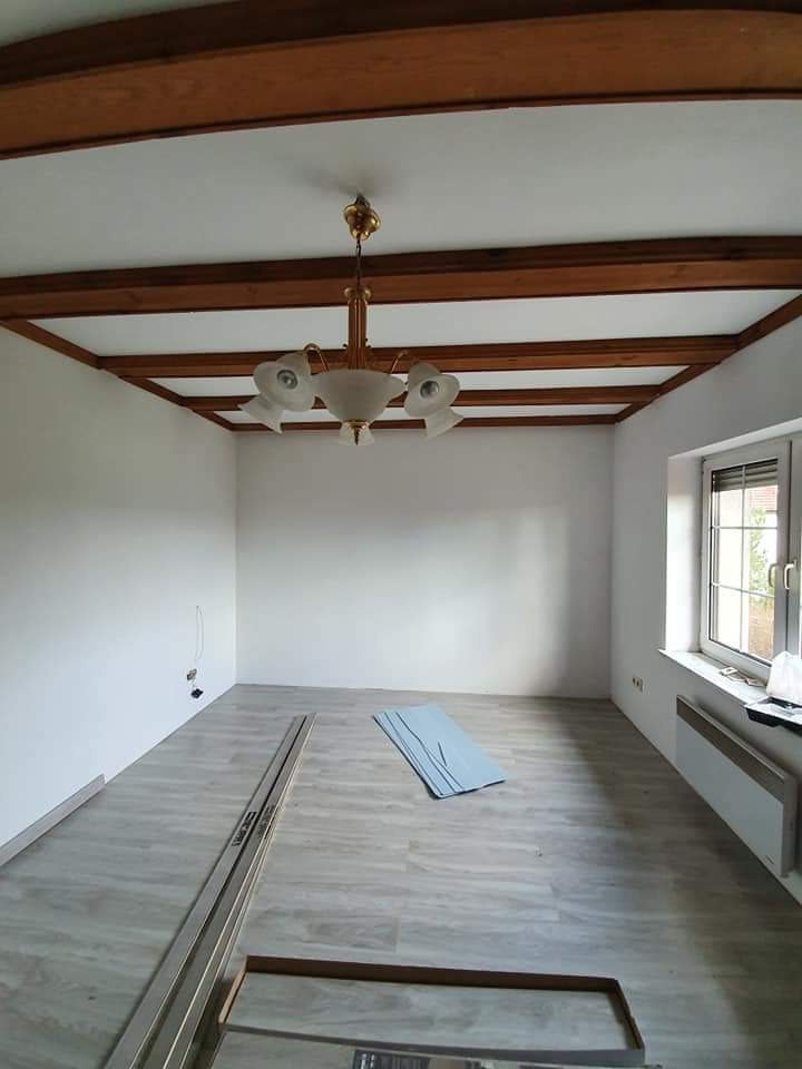 Pusty pokój po odświeżeniu z drewnianymi belkami na suficie, nową podłogą i świeżo pomalowanymi ścianami na biało, widoczny grzejnik pod oknem i elementy montażowe na podłodze.