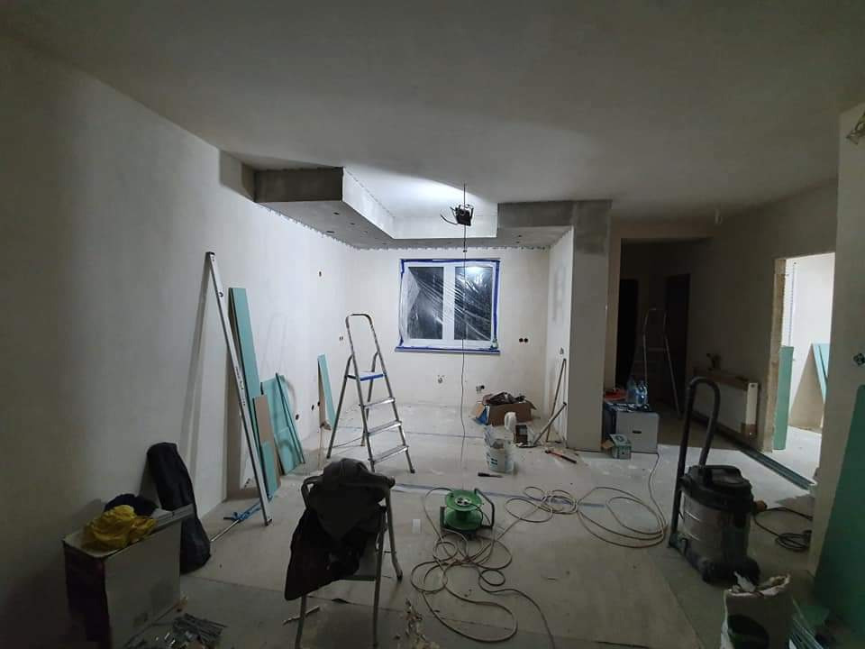 Wnętrze w trakcie remontu: widoczne płyty karton gips, drabina, narzędzia, zakryte folią okno i świeżo wygładzone ściany pod przyszłe malowanie.