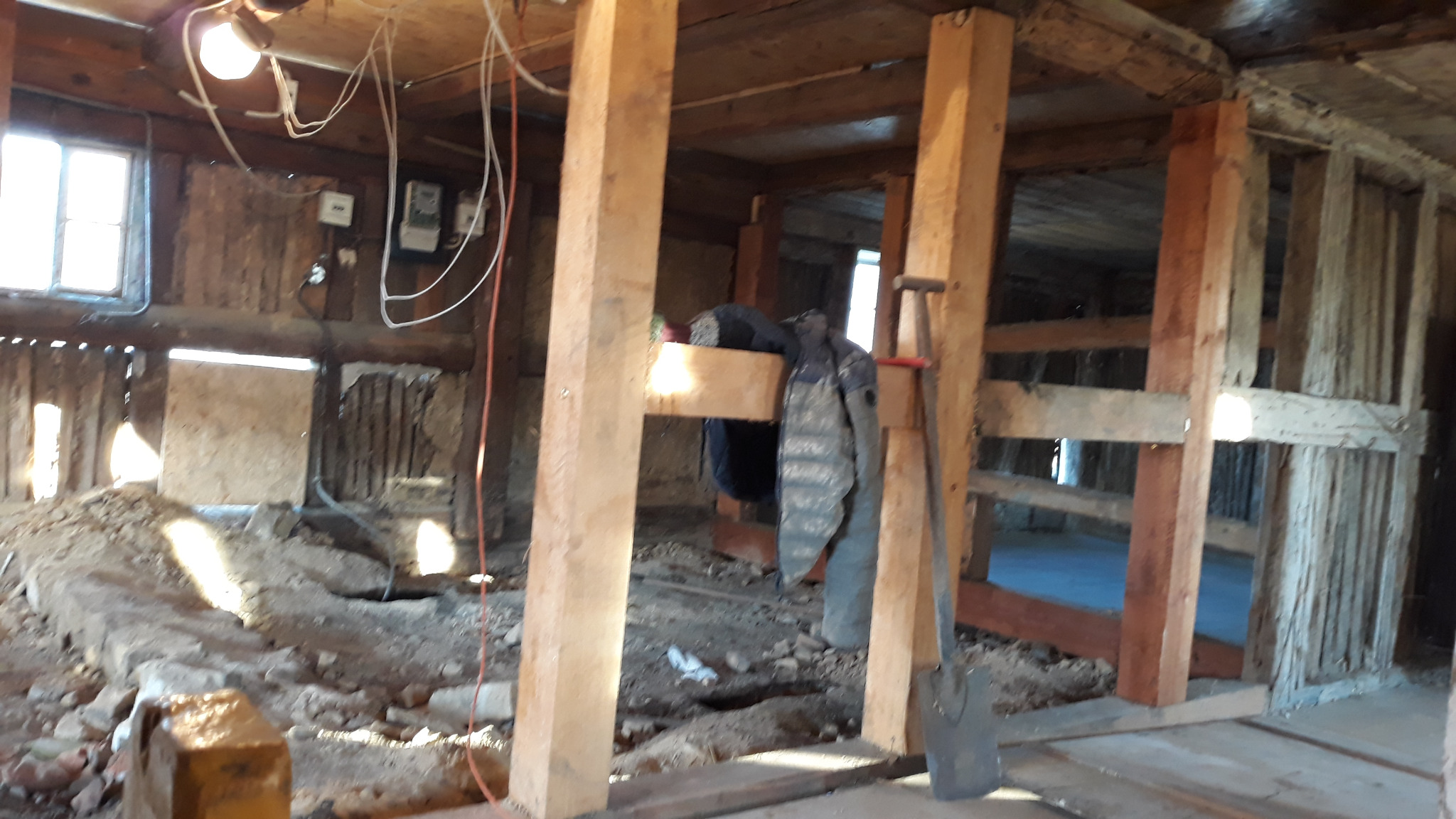 Wnętrze starego budynku w trakcie remontu, widoczne drewniane słupy wzmacniające konstrukcję, fragmenty ścian z odsłoniętą konstrukcją, ziemia na podłodze oraz narzędzia pracy, łopata oparta o słup.