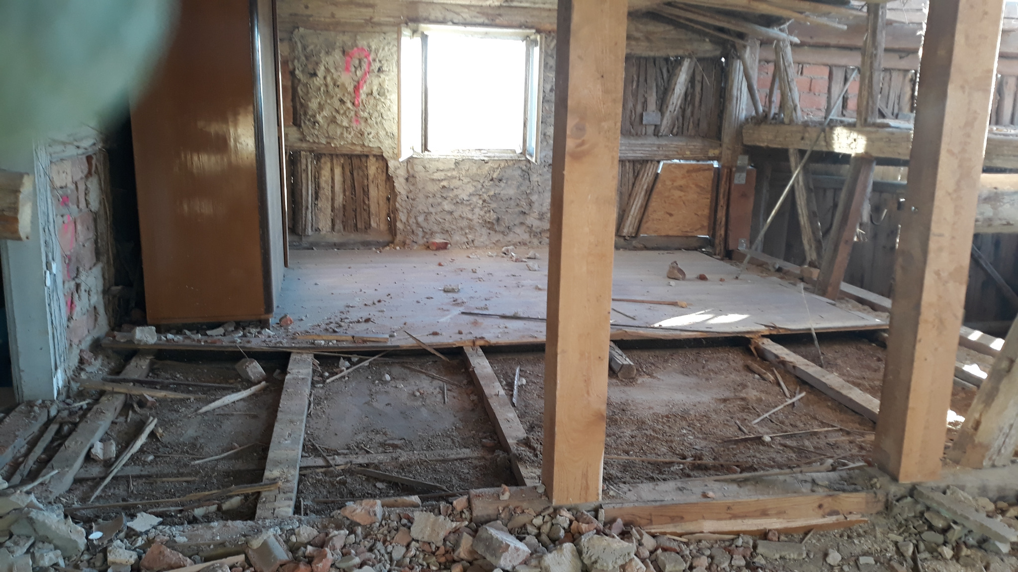 Wnętrze starego domu w trakcie rozbiórki, widoczne odsłonięte cegły, drewniane belki stropowe i fragmenty podłogi, pozostawione resztki gruzu i odpadów budowlanych.