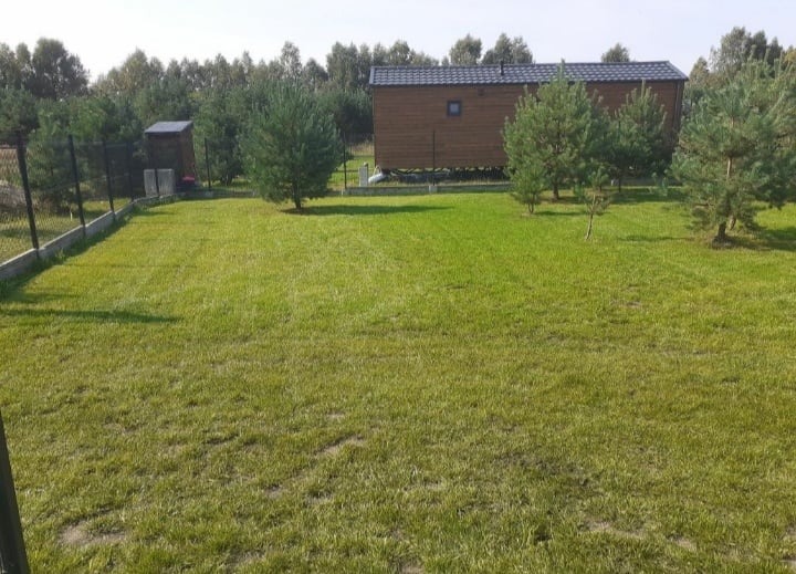 Starannie skoszony trawnik z młodymi drzewkami iglastymi, drewniany domek letniskowy na podwyższeniu w tle, ogrodzenie siatkowe z betonową podmurówką.