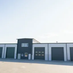 Nowoczesny budynek magazynowy z szarymi segmentowymi bramami garażowymi, oświetleniem zewnętrznym i kostką brukową przed budynkiem, w słoneczny dzień.