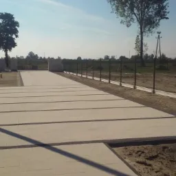 Wyłożona jasną kostką brukową powierzchnia z ciemnymi pasami, ogrodzona zieloną siatką z betonową podmurówką, w tle drzewa i błękitne niebo.