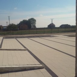 Modzelewski Budownictwo - Nowo ułożona, jasna nawierzchnia z kostki brukowej z ciemnymi pasami oddzielającymi sekcje, widok na budynek z oknem po lewej, słup betonowy w prawym dolnym rogu i krajobraz wiejski w tle...