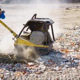 Operator zagęszcza gruz żółtą zagęszczarką wibracyjną; w tle unosi się pył.