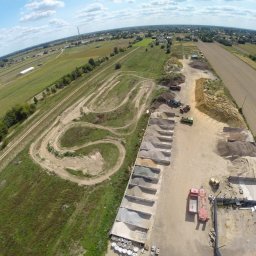Modzelewski Budownictwo - Widok z drona na plac z różnymi frakcjami kruszyw w betonowych przegrodach, obok tor ziemny z wyjeżdżonymi śladami pojazdów, w tle pola uprawne i zabudowania.