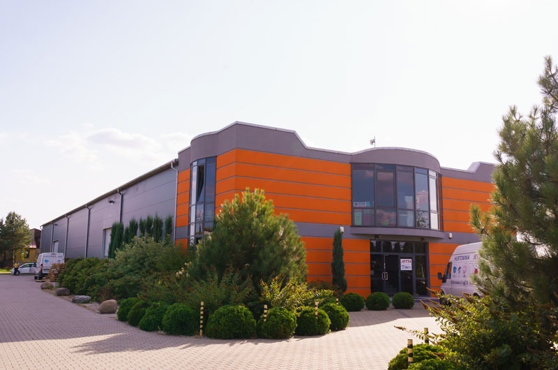 Nowoczesny budynek przemysłowy z pomarańczowo-szarą fasadą, otoczony zielenią i brukowanym chodnikiem, z zaparkowanymi samochodami dostawczymi w tle.
