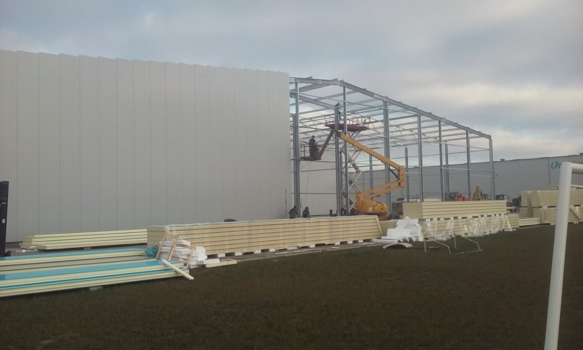 Montaż stalowej konstrukcji hali z wykorzystaniem podnośnika koszowego, widoczne płyty warstwowe i fragment elewacji.