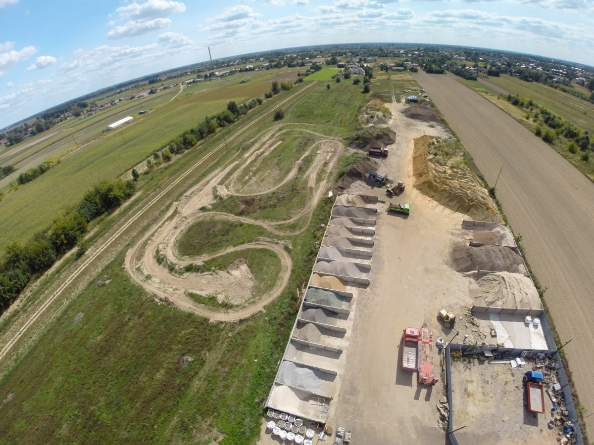 Widok z drona na plac z różnymi frakcjami kruszyw w betonowych przegrodach, obok tor ziemny z wyjeżdżonymi śladami pojazdów, w tle pola uprawne i zabudowania.