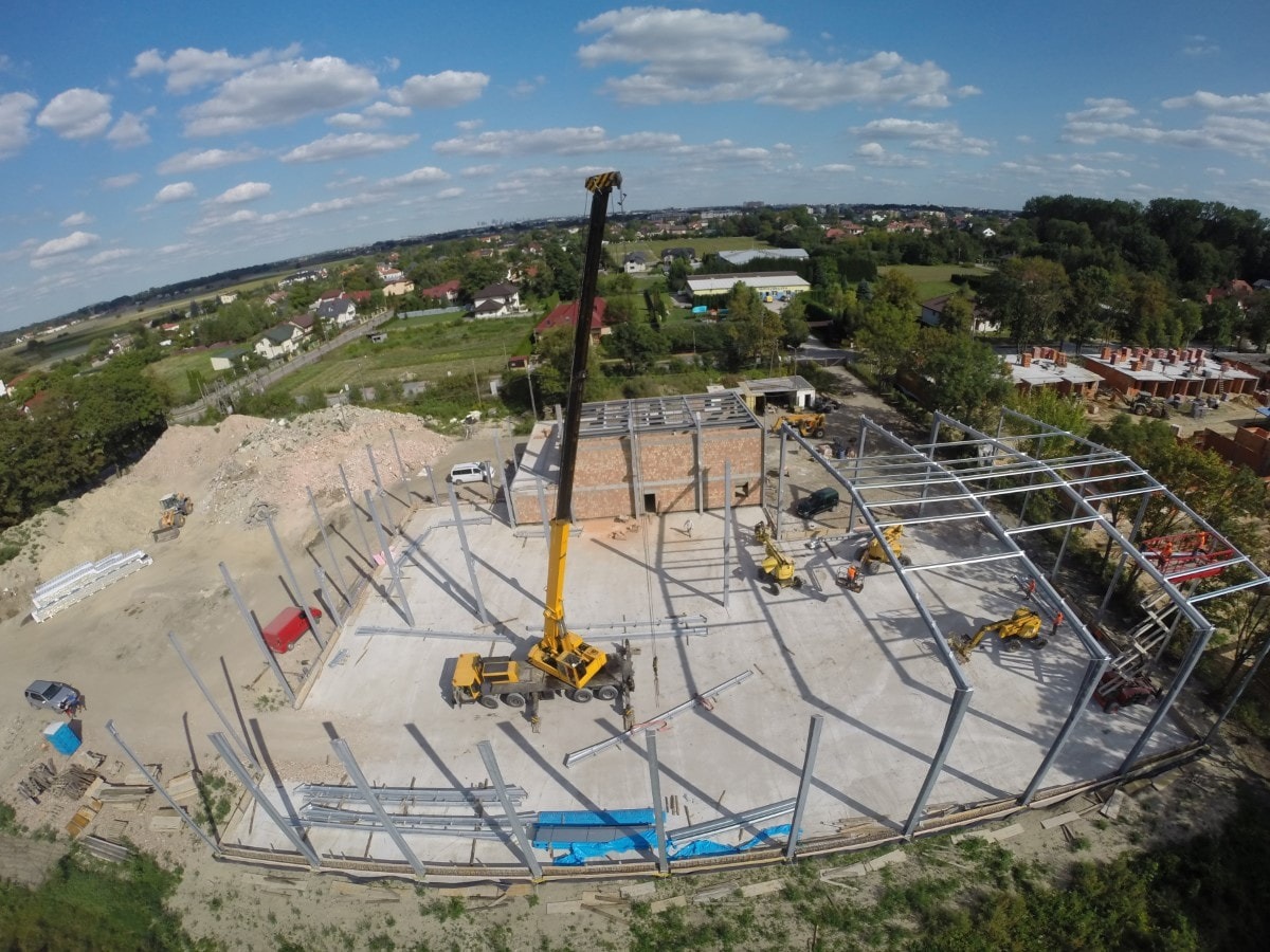 Szeroki kadr z lotu ptaka na plac budowy z rozstawioną stalową konstrukcją szkieletową budynku, żurawiem, kilkoma koparkami i stertą ziemi w tle.