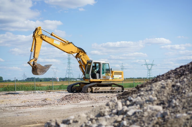 Żółta koparka na placu budowy w Płochocinie, z pryzmą gruzu w pierwszym planie i słupami wysokiego napięcia w tle, podczas prac ziemnych.