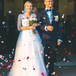 Uśmiechnięta para młoda opuszcza kościół obsypywana płatkami róż; panna młoda w białej sukni z bukietem białych róż, pan młody w granatowym garniturze z muchą i okularach.