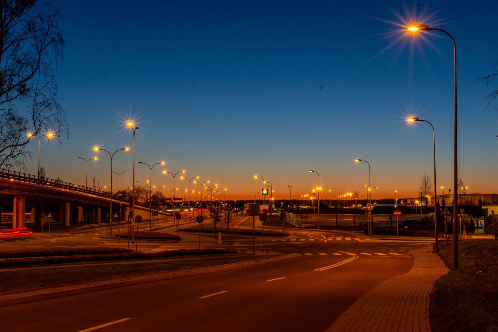 Miejski krajobraz o zmierzchu: skrzyżowanie z licznymi latarniami, widoczne smugi świetlne samochodów i wiadukt w tle.