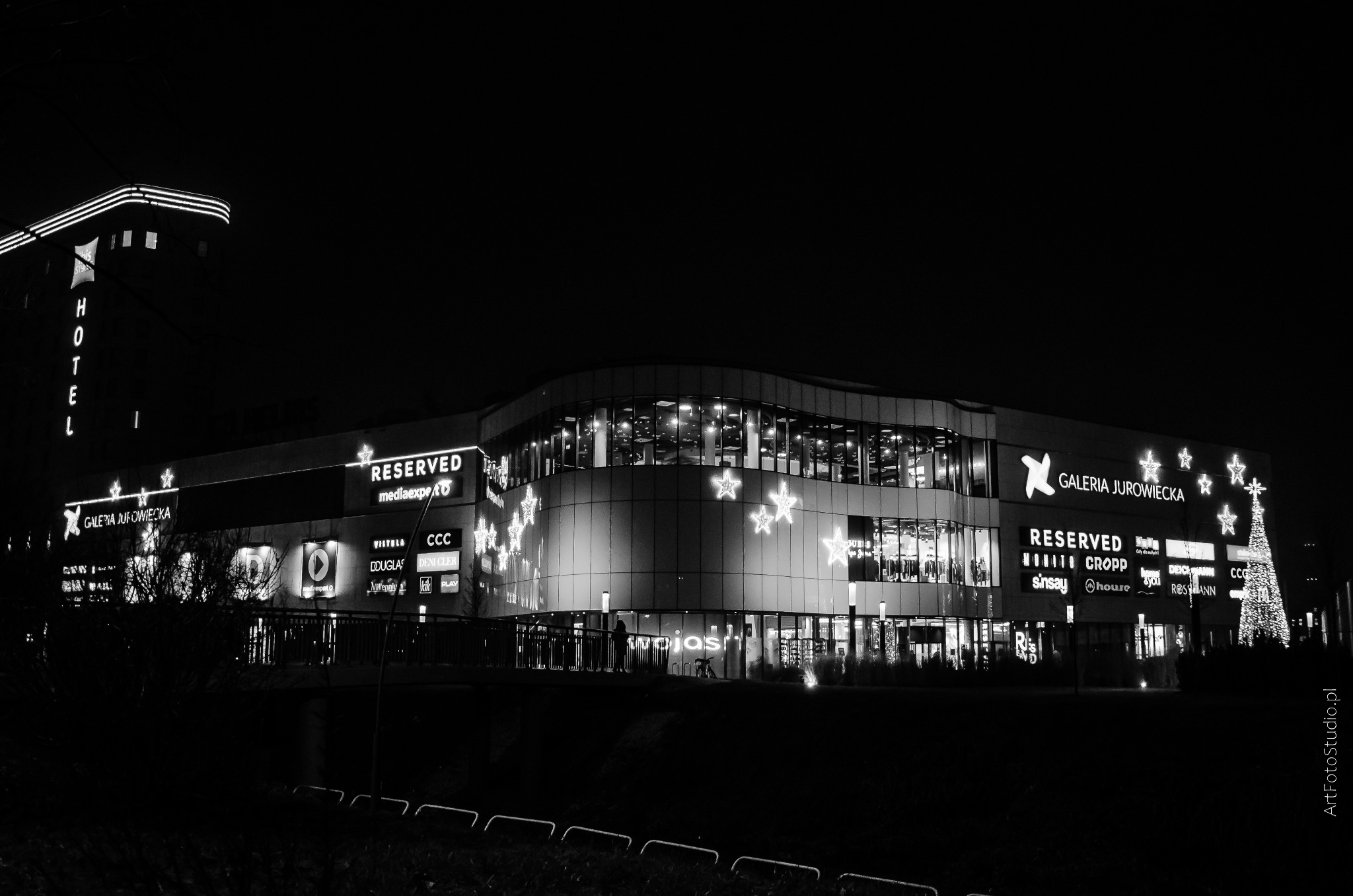 Czarno-białe zdjęcie nocne oświetlonej Galerii Jurowieckiej z hotelem w lewym górnym rogu, udekorowanej świątecznymi gwiazdami i choinką z świateł.