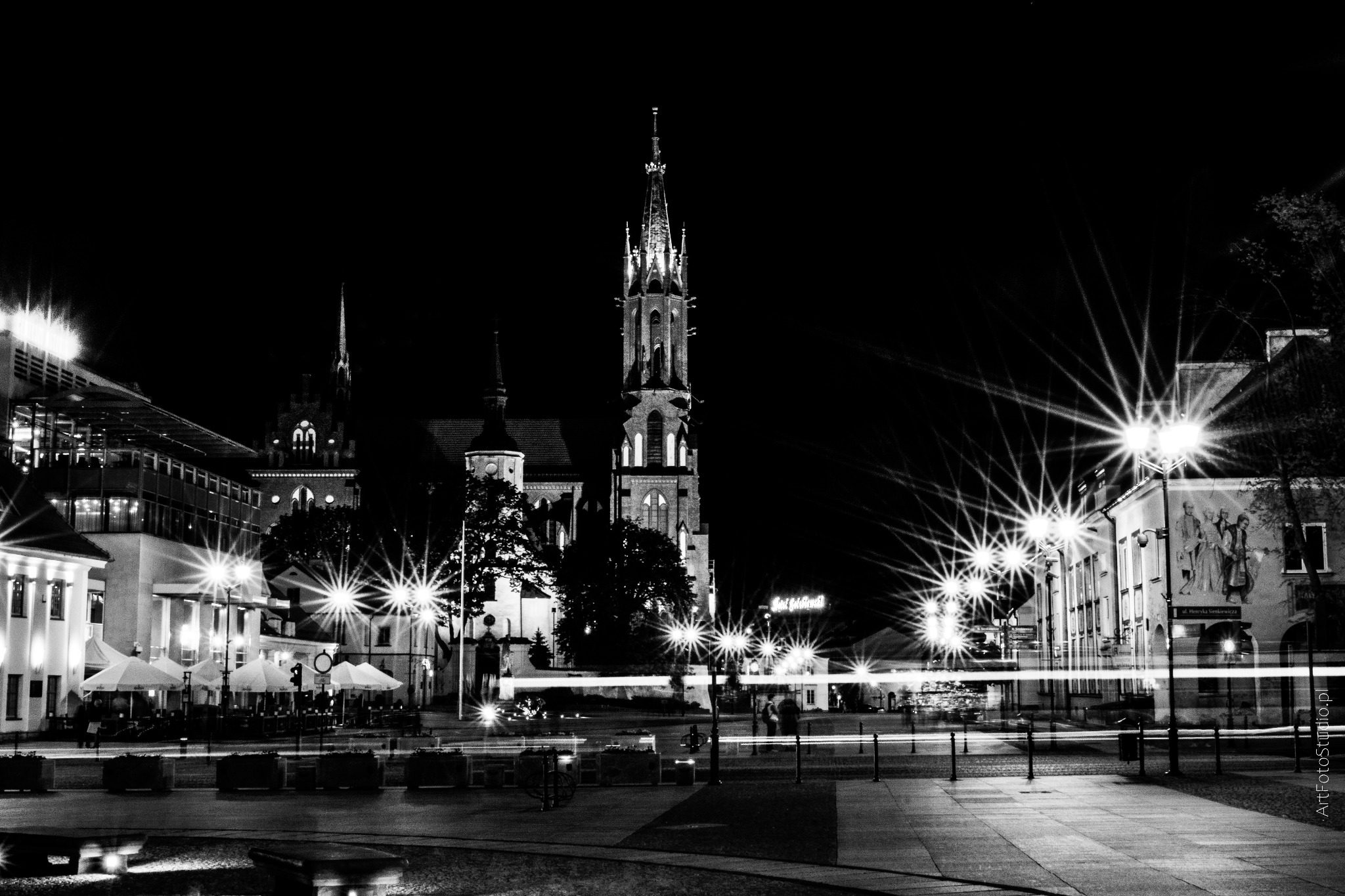 Nocny widok na oświetloną gotycką katedrę i budynki miejskie w czerni i bieli, z efektem smug świateł samochodów.