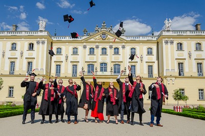 Grupa absolwentów w togach i biretach rzuca czapkami w powietrze na tle pałacu z elementami barokowymi w słoneczny dzień.