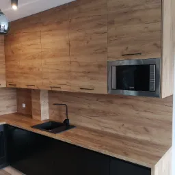 Nowoczesna kuchnia z frontami w kolorze drewna, czarnym blatem i minimalistyczną armaturą. W tle widać wbudowaną kuchenkę mikrofalową w srebrnej obudowie i designerską lampę wiszącą.