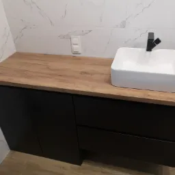 Nowoczesna łazienka: czarna szafka pod umywalkę z blatem imitującym drewno, biała umywalka nablatowa i czarna bateria na tle marmurowych płytek ściennych.