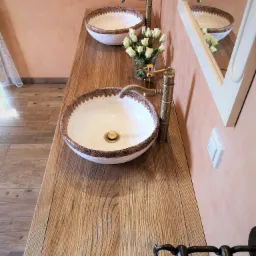 Dwie ceramiczne umywalki nablatowe z dekoracyjnym brzegiem i stylizowanymi kranami na blacie z litego drewna o widocznej strukturze słojów. W tle wazonik z białymi różami i fragment lustra...