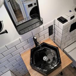 MAGURA - Czarna, sześciokątna umywalka nablatowa z czarną baterią, ustawiona na blacie z jasnego drewna, w nowoczesnej łazience z białymi płytkami imitującymi cegłę. W lustrze odbija się pralka i torba...