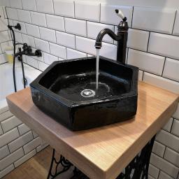 MAGURA - Czarna, ośmiokątna umywalka nablatowa z chromowanym odpływem i czarną baterią, zamontowana na drewnianym blacie wspartym na metalowej konstrukcji maszyny do szycia, w łazience z białymi płytkami...